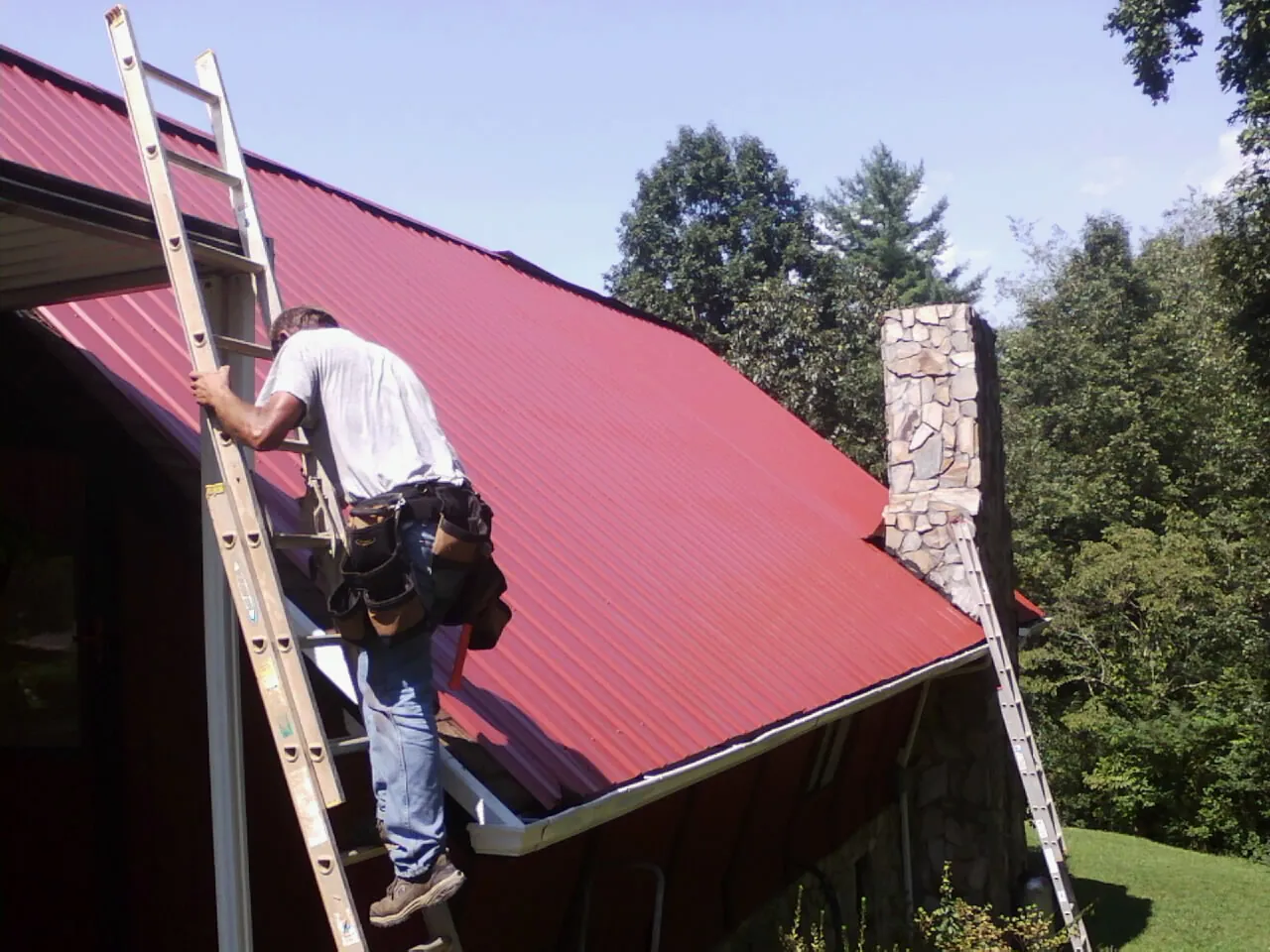 Roofer on a ladder beside a red metal roof and stone chimney — Soffit & Fascia Repair by Roofing Brothers in Barataria